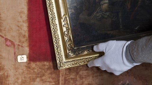 A gloved hand lifting a painting off of a wall to reveal the original dark red wallpaper colour behind it